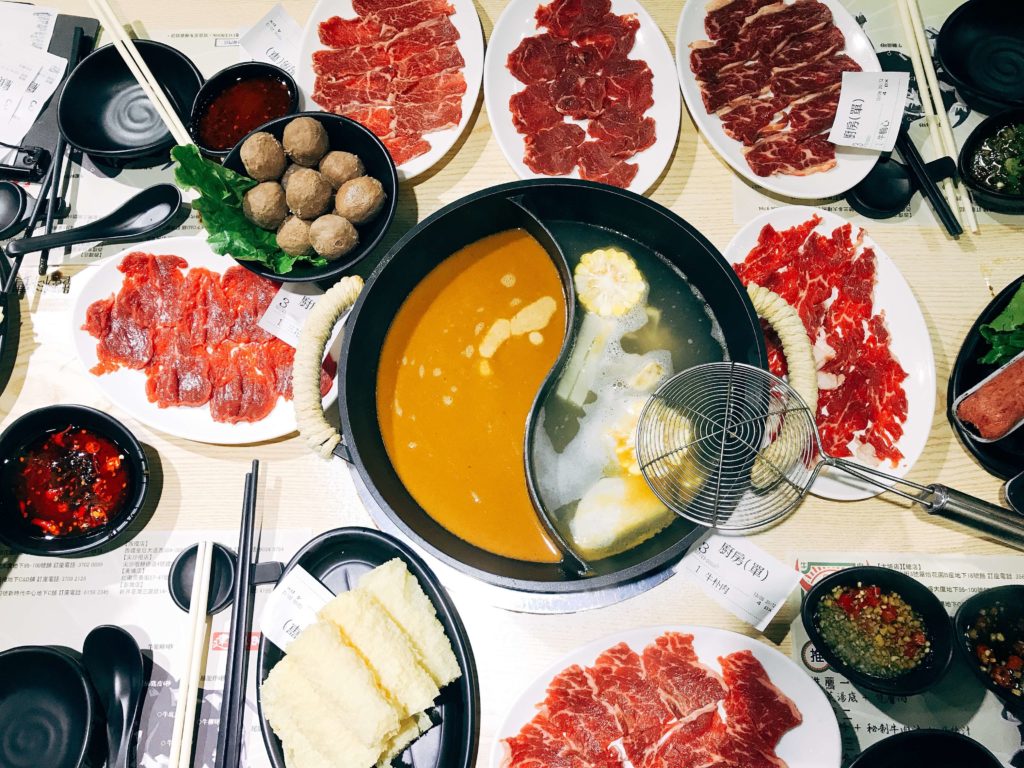 Traditional Chinese hot pot setup with yin-yang broth, surrounded by raw beef, lamb, shrimp paste, tofu skin, leafy greens, and dipping sauces, arranged for communal dining.