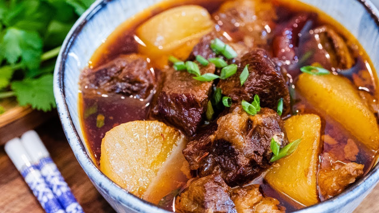 Bowl of braised beef brisket with daikon in rich soy-based broth, garnished with green onions, served with patterned chopsticks and fresh cilan