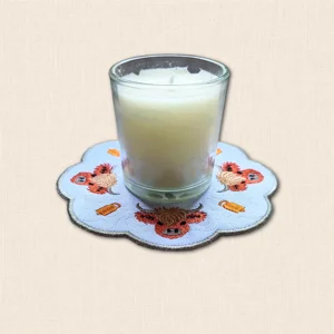 Highland Cow embroidered candle mat with glass candle jar, showing rustic cow design and scalloped edge