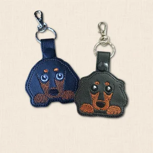 Two embroidered Kawaii Dachshund keyrings in black faux leather, showing both sausage dog designs with metal keyring and lobster clasp
