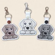 Three embroidered Labrador keyrings in Black, Chocolate and Golden Labrador designs, shown side by side on white faux leather