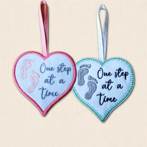 One Step at a Time heart hanger in two colour and stitch finishes, featuring embroidered footprints, encouraging wording and ribbon loops.