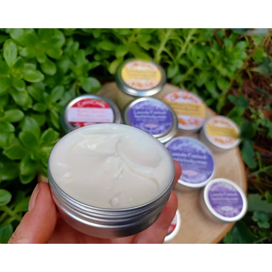 A round silver metal tin filled with 60g thick white hand and body cream. The lid is removed, revealing the smooth texture of the cream. Several other closed silver tins with colourful labels are arranged in the background.