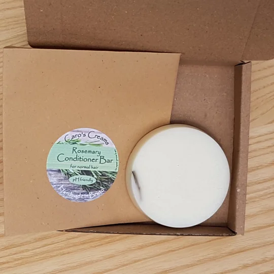 A round, white conditioner bar in a cardboard box with a label that reads "Caro's Creams Rosemary Conditioner Bar for normal hair pH friendly Handmade in Bristol", with a kraft envelope. The bar contains strands of rosemary, and is suitable for frequently washed hair