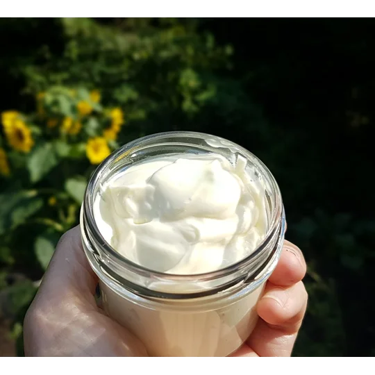 A clear glass jar filled with a thick, smooth, off-white cream. The substance has a soft, whipped texture with visible peaks and swirls on the surface, contained within a simple, round, transparent vessel with a wide opening.