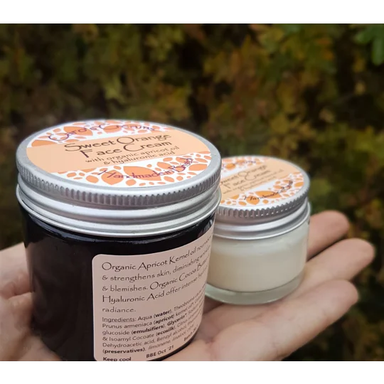 Two round glass jars of Sweet Orange Face Cream with silver metal lids. The larger jar is 60g features an orange label with orange leaf graphics and text detailing ingredients like organic apricot oil and hyaluronic acid. The smaller 20g glass jar is transparent, with an aluminium lid. This cream is suitable for all skin types.