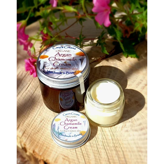 A small transparent glass jar of 20g of Caro's Creams Organic Argan Chamomile Cream for dry and mature skin, handmade in Bristol. The jar has a silver lid with a label featuring chamomile flowers. An open jar shows the pale yellow cream inside. There is an amber glass jar for the 60g option.