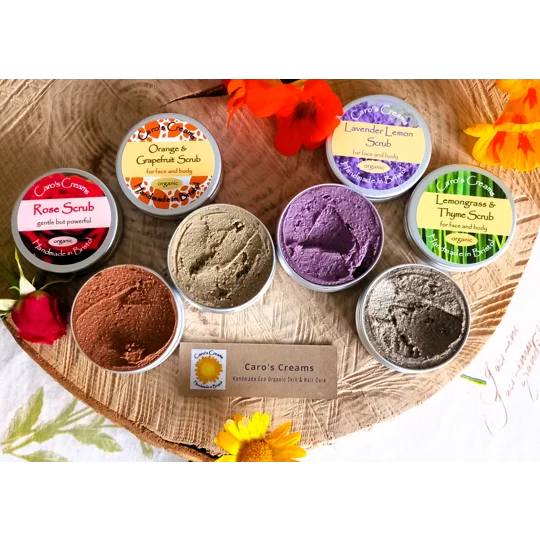 Four round metal tins of Caro's Creams body scrubs in Rose, Orange & Grapefruit, Lavender Lemon, and Lemongrass & Thyme, each with a label. Four open containers reveal textured scrubs in shades of red, brown, purple, and grey.