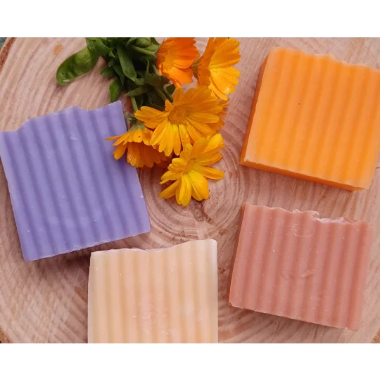 Four rectangular bars of soap with a ribbed texture in shades of purple, bright orange, pale peach, and dusty rose, arranged around a small cluster of fresh, vibrant orange calendula flowers with green stems and leaves.