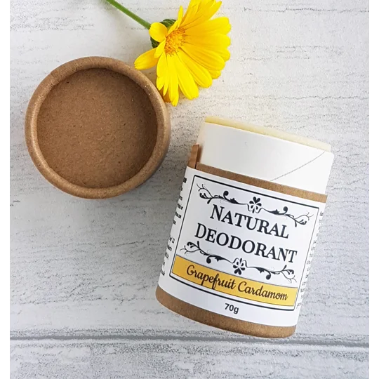 A cylindrical cardboard tube of natural deodorant with a cream-coloured solid deodorant inside. The white label features black text reading Natural Deodorant and Grapefruit Cardamom with a yellow accent bar and the weight listed as 70g.