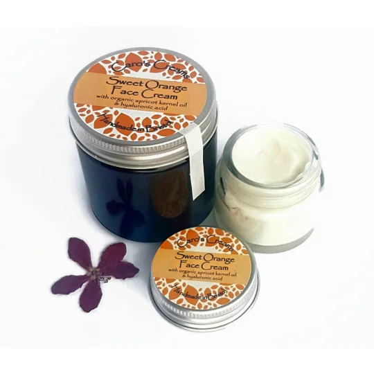 An amber glass jar of 60g Caro's Creams Sweet Orange Face Cream with organic apricot kernel oil and hyaluronic acid, alongside an open clear glass jar containing 20g off-white cream and a separate matching lid, plus a small purple flower.