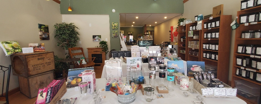 the inside of the tearoom with a table covered in tea related gifts. in the background there are shelves covered in pacaged tea and the cozy corner with antique tea shipping crates a rocking chair and a fire place.
