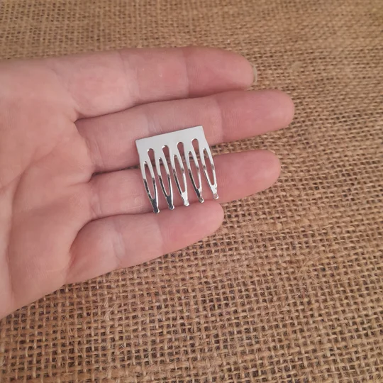 A hand holding a small silver metal doll hair comb with five tines against a brown burlap fabric background, demonstrating the micro scale of collectible doll hair accessories.