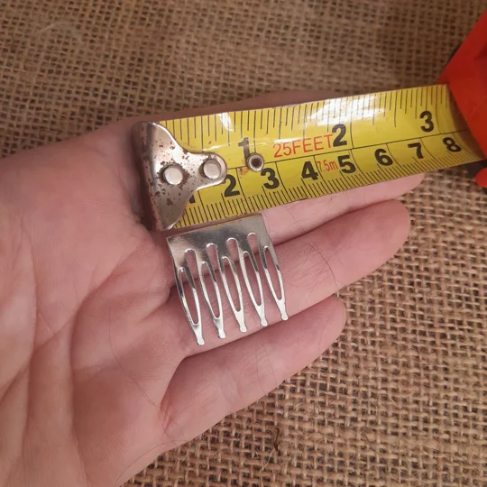 Silver metal miniature hair comb for dolls held in a hand over a burlap surface.