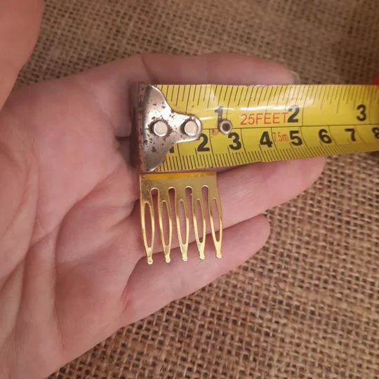 Gold metal miniature doll hair comb blank held in a hand for scale comparison against a burlap fabric surface.