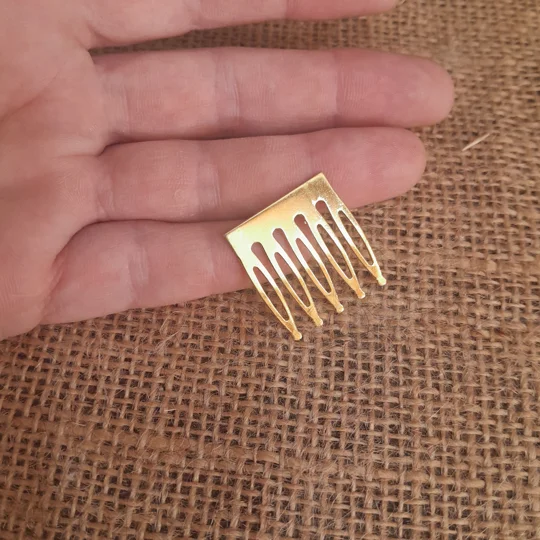 A person holding a miniature gold metal doll hair comb with five prongs over a rustic brown burlap background to showcase the micro scale of adult collector doll accessories.
