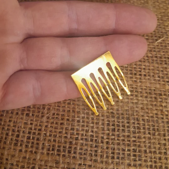 A miniature six tine gold metal hair comb for dolls, shown in a hand, ideal for custom collector doll wig styling and restoration.
