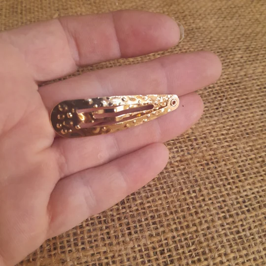 A person holding a polka dot gold metal doll hair clip or miniature snap clip against a brown burlap background to illustrate the micro scale for adult collector dolls.
