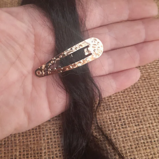 A miniature polka dot gold metal snap clip blank shown open and gripping dark doll hair for scale, used for adult collector doll hair accessories and wig styling.