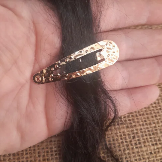 Gold textured miniature snap clip for dolls shown open and secured onto a strand of dark suri doll hair held in a hand.
