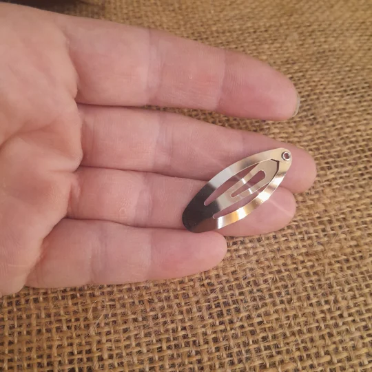 A hand holding a classic silver metal snap barrette for dolls against a brown burlap background to illustrate the micro scale of custom adult collector accessories.