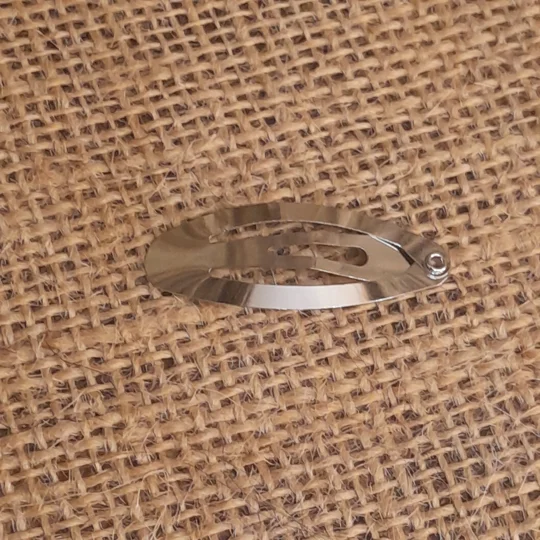 A small silver oval snap clip or barrette blank placed on a textured burlap surface, perfect for crafting custom hair pieces for suri doll hair.