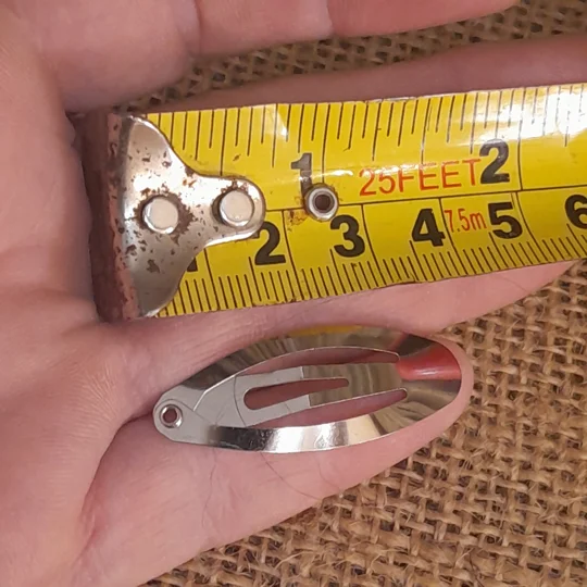 A silver metal doll snap barrette shown next to a yellow measuring tape to display its micro scale size of approximately three centimeters.