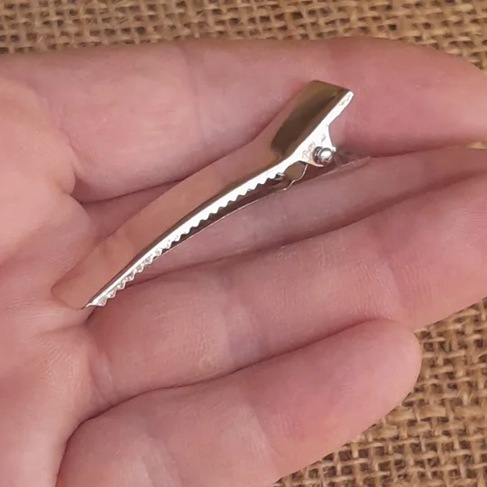 A hand holding a miniature silver metal alligator hair clip for dolls, viewed from the side profile to show the spring mechanism and serrated teeth.