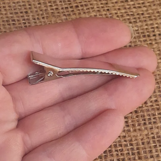 A hand holding a miniature silver metal alligator hair clip for dolls, viewed from the side profile to show the spring mechanism and serrated teeth.