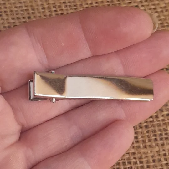 The top view of a small silver rectangle alligator clip blank held in a hand against a natural brown burlap background for scale.