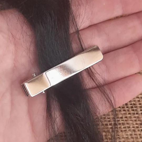 A miniature silver metal alligator clip fastened onto a lock of dark suri doll hair to demonstrate sizing for adult collector doll hair styling.
