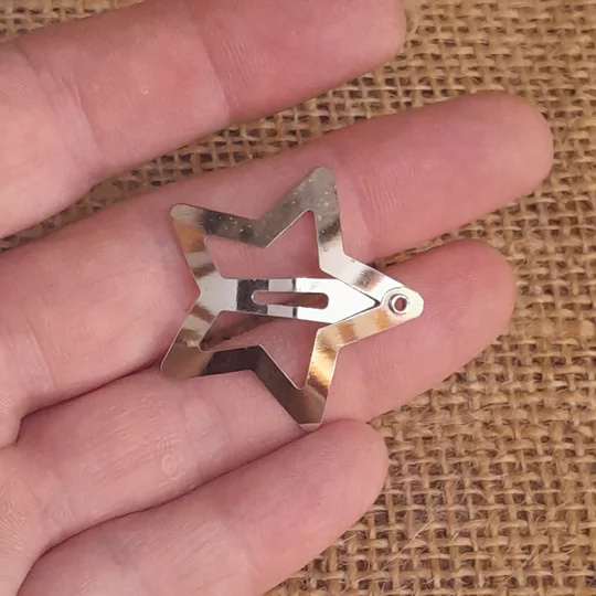 A hand holding a blank silver metal star shaped doll snap clip showing the front side mechanism against a rustic brown burlap background for scale.