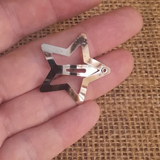 A micro scale silver star hair clip for dolls held open in a hand, showing the back snap clip prong used for securing hairstyles on collectible dolls.