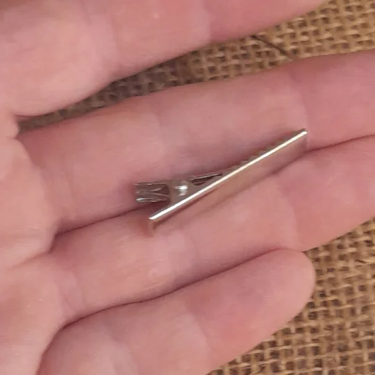 A hand holding a tiny silver metal alligator clip for dolls, viewed at an angle to showcase the spring hinge configuration