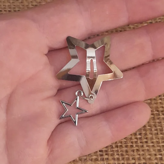 A person holding a silver metal star shaped doll snap clip featuring a small dangling open star charm on a delicate chain against a brown burlap background to demonstrate micro scale styling