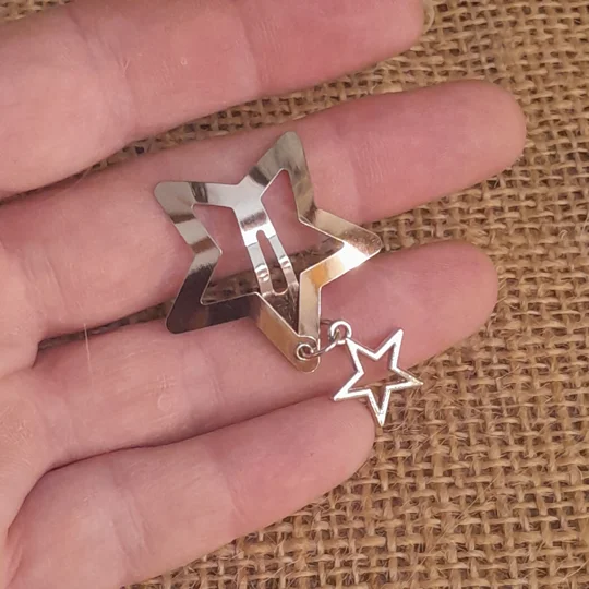 A person holding a silver metal star shaped doll snap clip featuring a small dangling open star charm on a delicate chain against a brown burlap background to demonstrate micro scale styling.