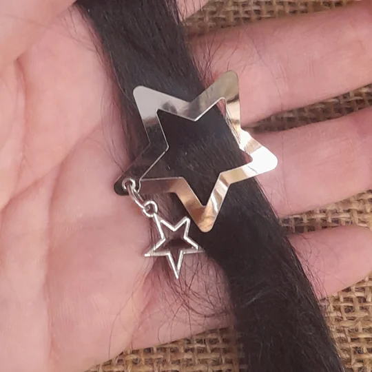 Silver star shaped miniature doll hair clip with a dangling star charm shown open and fastened onto a strand of dark suri doll wig hair held in a hand.