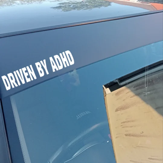 close up view of a car windscreen with a matte black sunstrip with driven by adhd text sticker offset left in picture in colour white