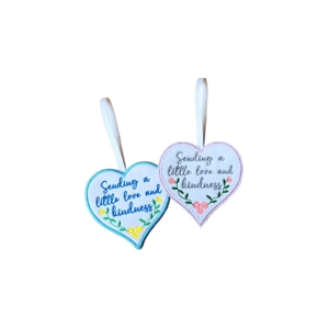 Two handmade felt heart keepsakes with hanging ribbons, embroidered with the words ‘Sending a little love and kindness’. One has a turquoise border with blue text and yellow flowers, the other a lilac border with grey text and pink flowers.