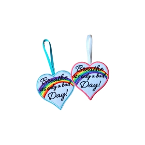 Two felt heart-shaped hanging keepsakes embroidered with the words “Breathe – it’s only a bad day!” and a rainbow across the centre. One heart has a turquoise stitched edge with a matching hanging loop, and the other has a pink stitched edge with a white hanging loop.