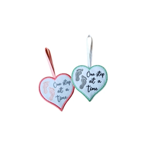 Two felt heart decorations embroidered with small footprints and the words ‘One step at a time’, each finished with a hanging ribbon.