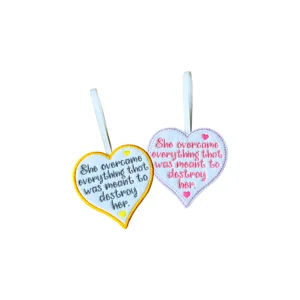 Two white felt heart decorations with embroidered quote “She overcame everything that was meant to destroy her”, shown in grey and pink text versions with yellow and lilac stitched borders, small heart details and white hanging loops