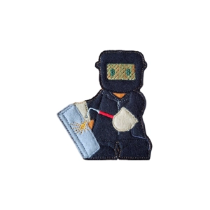 A felt gingerbread welder character wearing protective overalls and a welding mask, holding a welding torch beside a metal plate with stitched sparks.