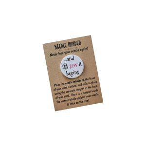 A kraft-brown backing card labelled 'Needle Minder – Never lose your needle again!' holds a round white needle minder. The minder shows a small spool illustration and the phrase '...and sew it begins', with the word 'sew' in pink. Text on the card explains how to place the needle minder on fabric using the magnet on the back.