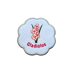 A scalloped candle coaster with an embroidered pink gladiolus flower and green leaves on a white background, with the word “Gladiolus” stitched below the flower.