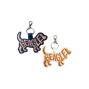Beagle-shaped faux leather keyrings featuring tan embroidered word-art, one in black and one in white, each with a metal keyring and clasp.