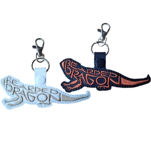 Two embroidered Bearded Dragon keyrings shown side by side, one in black with orange lettering and one in white with beige lettering. Each keyring features the words “Bearded Dragon” stitched to form the shape of the reptile, finished with a metal clasp and keyring attachment.