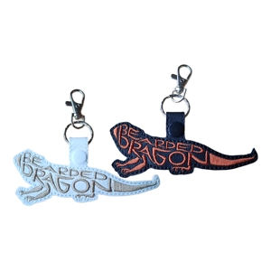 Two Bearded Dragon Word Art Keyrings, one in white with beige embroidery and one in black with dark copper embroidery. Each keyring is shaped like a bearded dragon, with the words 'Bearded Dragon' forming the body in stitched word art. Both have a snap tab and metal clip attached.