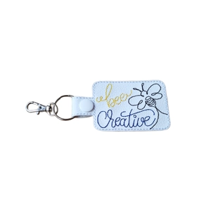 Bee Creative faux leather keyring, machine embroidered with the words ‘Bee Creative’ in yellow and blue text and a bee illustration, finished with a metal keyring and lobster clasp.