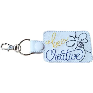 Bee Creative embroidered faux leather keyring in white, with bee design, yellow and black stitched lettering and metal clip.